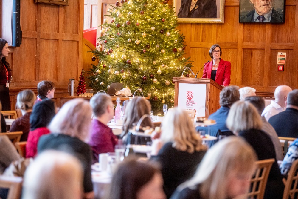 large group of seated people listening to an address being given by a women in red jacket standing beside a Christmas tree in an old, indoor event space