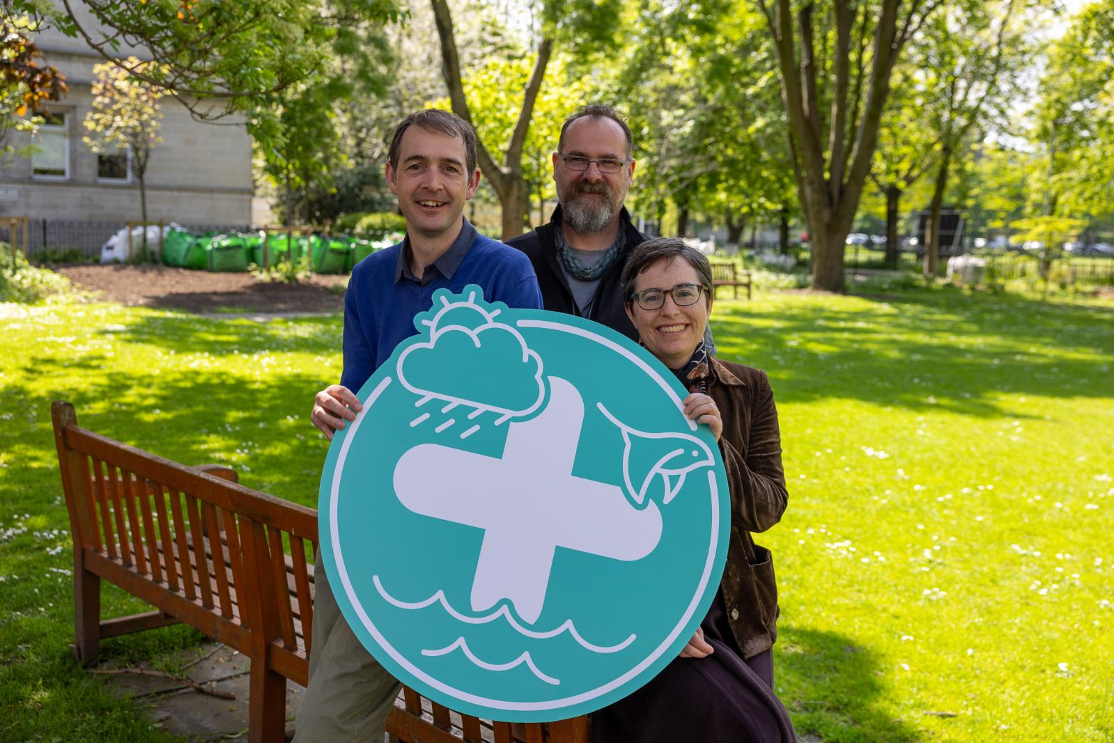 Two men and one woman stand in a garden holding a teal logo sign.