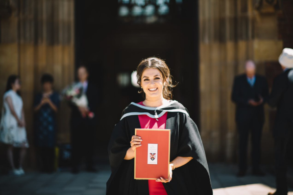 female student standing outside Queen's University main door in graduation gown