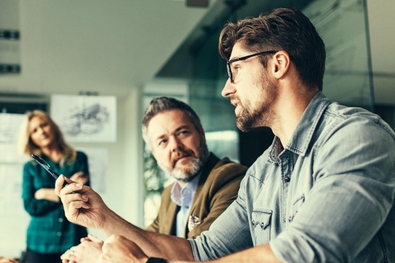 Microsoft Office background of a breakout session showing seated man with glasses talking and being listened to by a standing woman and another seated man