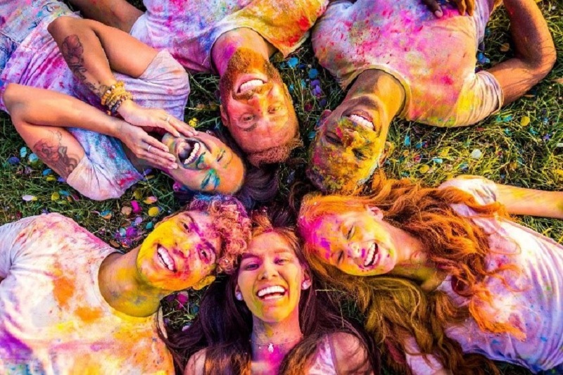 smiling young people covered in coloured powders lying in a circle with their heads together