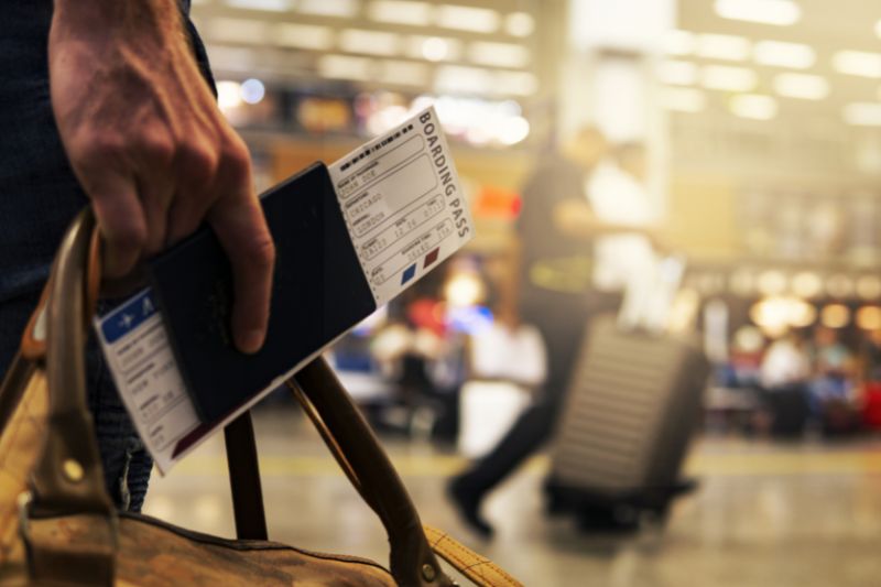 Hand holding a boarding pass insie a passport against a brightly lit airport backdrop with people walking in the distance with suitcases.