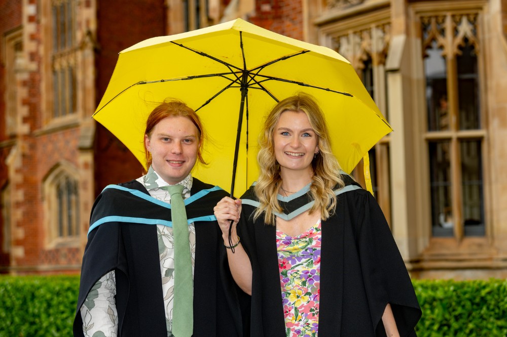 male and female graduates smiling under a yellow umbrella