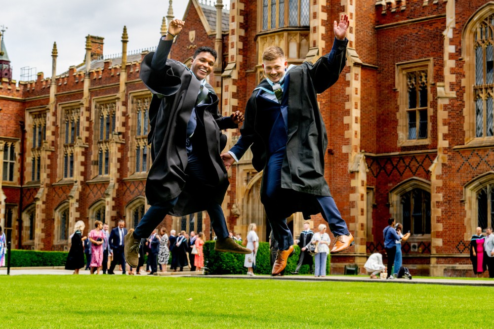 two male graduates jumping in the air in celebration on Queen's University Belfast front lawn