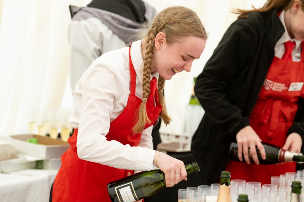 two young female student catering staff pouring sparkling refreshment into glasses in the graduation marquee