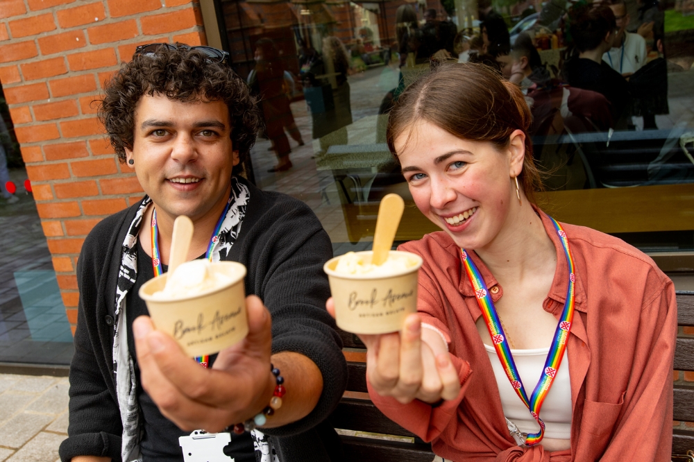 two smiling people with rainbow lanyards holding up their ice cream outside the Main Site Tower at the QUB Pride Picnic 2023