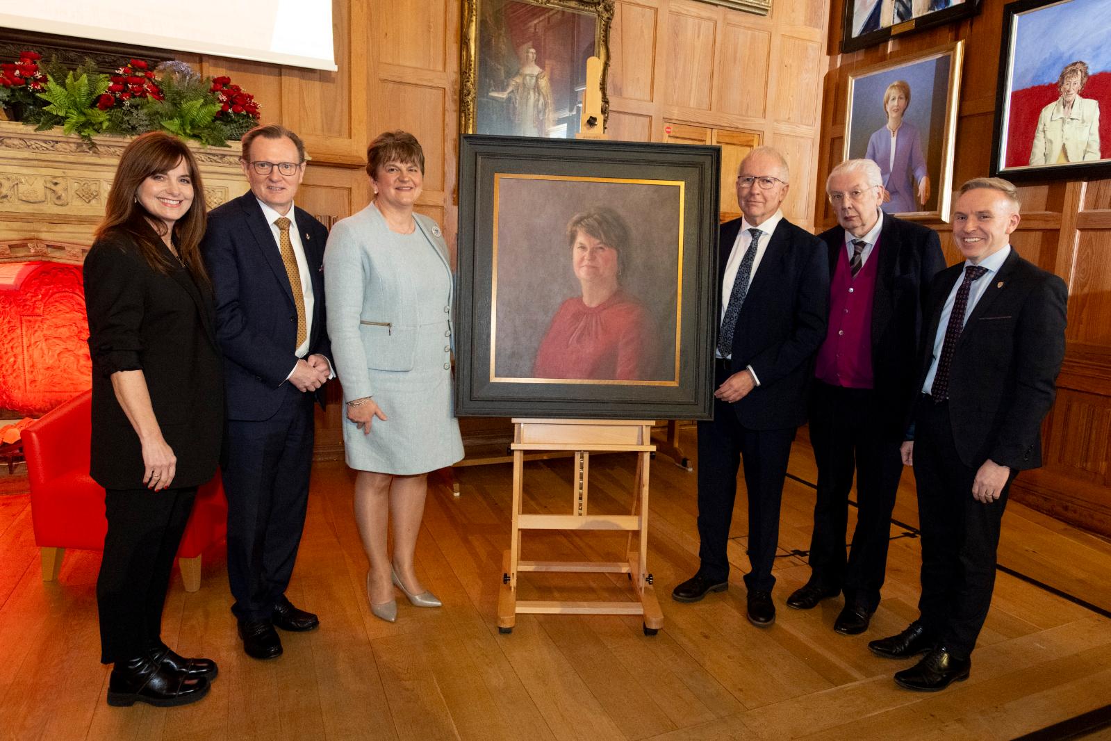 A group of people stand posed beside a portrait in a university grand hall, looking towards the camera.