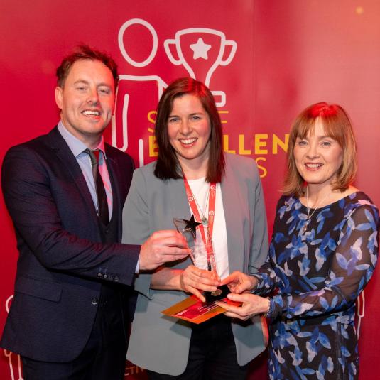 A man and two women standing in front of a large red Staff Excellence Awards banner. Lady in the middle is holding a glass trophy award