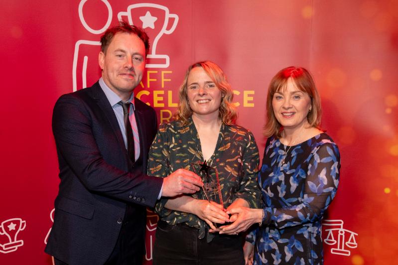 A man and two women standing in front of a large red Staff Excellence Awards banner. Lady in the middle is hold a glass trophy award
