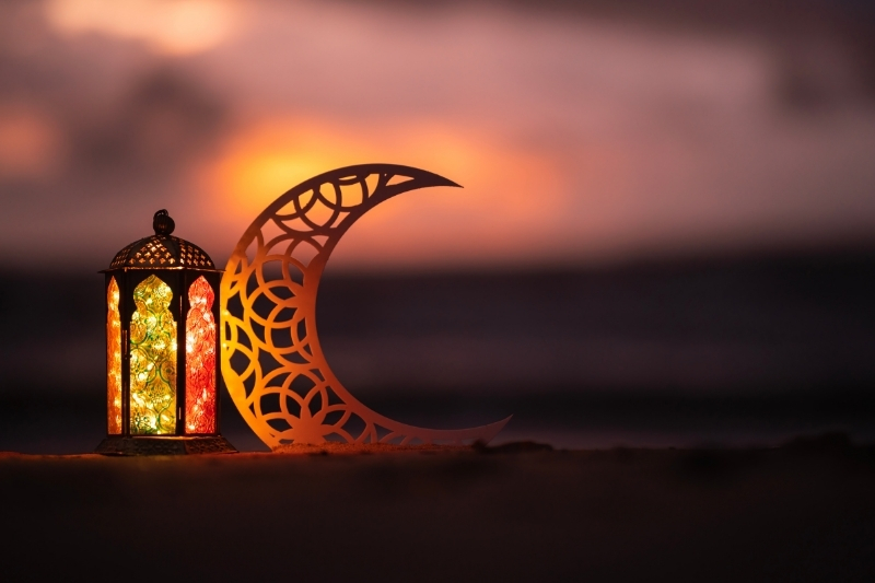 A decorative lantern with intricate patterns and warm glowing lights stands beside an ornate crescent moon cutout on a sandy surface at sunset. The background features a blurred sky with hues of orange, pink, and purple.