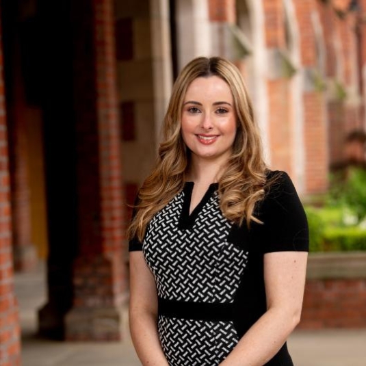 young woman in black and white patterned dress smiling to camera in a cloistered space with greenery in the background