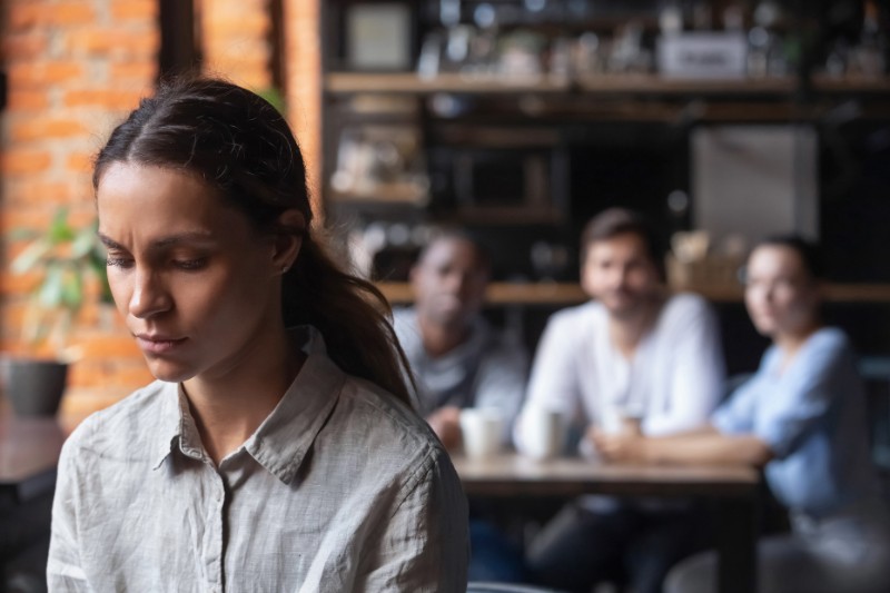 sad and anxious looking young woman standing and looking at the ground with blurred, seated group of people looking at her in the background