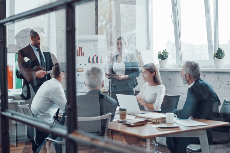 four men and two women in business-wear, seen through a glass panel, in discussion at a table in a meeting room