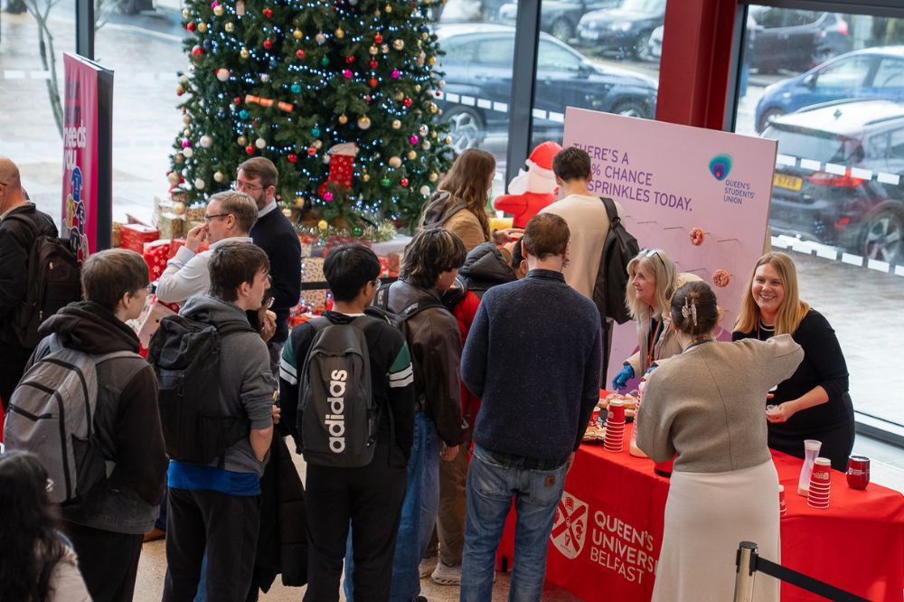 A crowd of students gathers around a Queen’s University Belfast stall offering festive treats, with a decorated Christmas tree behind the