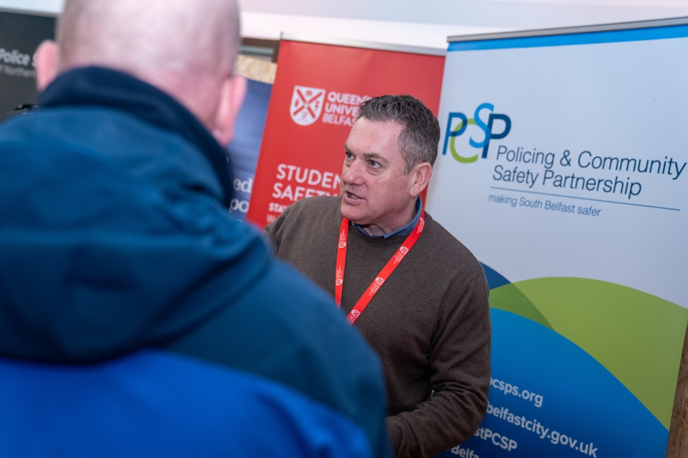 A representative speaks with an attendee at a campus safety information stall, with University and community safety banners behind them.
