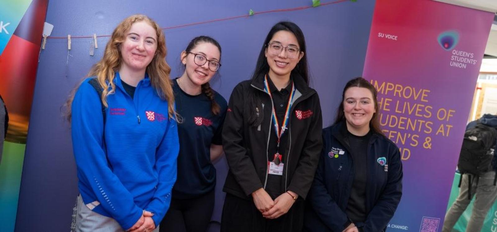 four young women smiling to camera while standing at a promotional stand at an indoor event