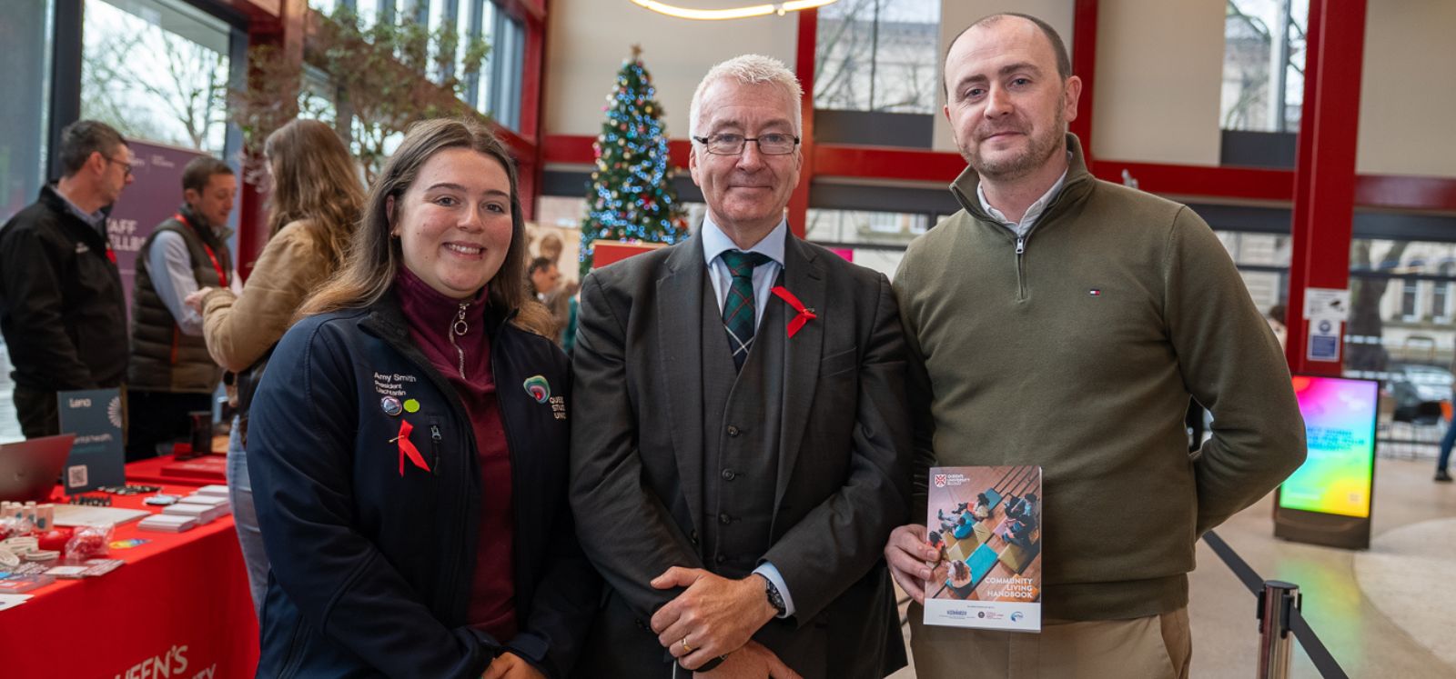 Three people stand together at an indoor event, smiling at the camera. A decorated Christmas tree and information tables are visible in the background. One person holds a booklet, and all three wear red awareness ribbons.
