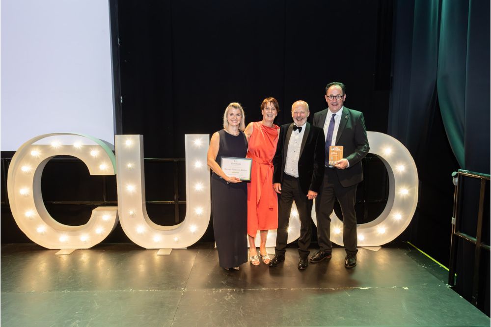 Two male and two females standing in front large lightbox CUBO letters holding an award and certificate