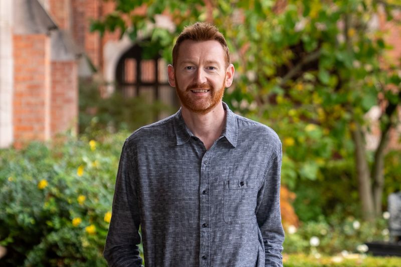 Alt text: Portrait of a man standing outdoors on a university campus, smiling at the camera, with brick buildings and greenery in the background.