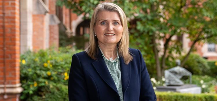 smiling blond-haired woman wearing navy-blue jacket pictured in a courtyard against a background of green foliage