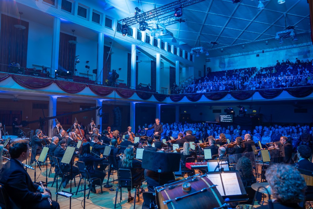 a large blue-lit hall showing an orchestra in the fore and midground and seated audience listening on ground and balcony floors