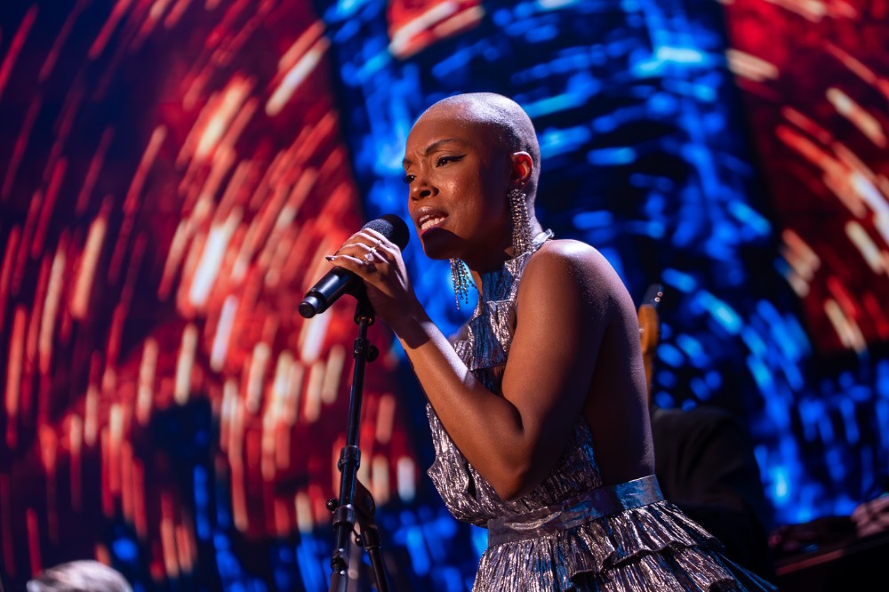 black woman singing into a microphone on stage with a swirling pattern showing on a background presentation screen