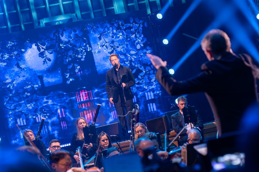 man singing on a raised blue-lit stage with orchestra in the foreground