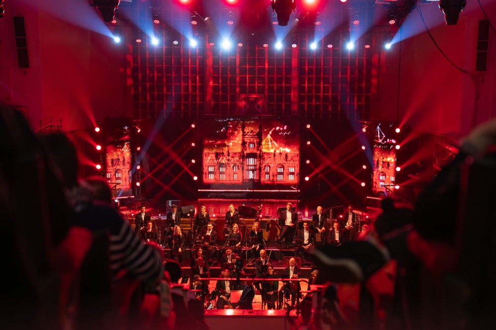 long view from the balcony of a large hall showing a seated audience in the foreground and seated orchestra on stage in the background against a presentation screen