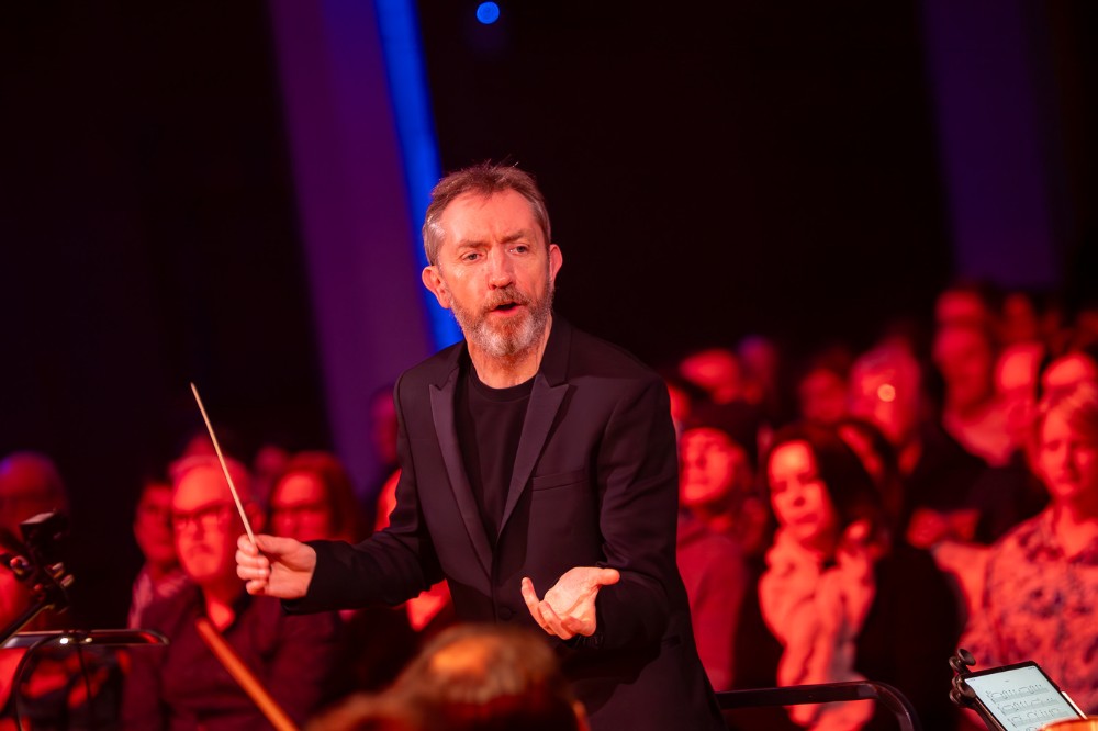 orchestral conductor waving his baton at a concert, with audience pictured in the background