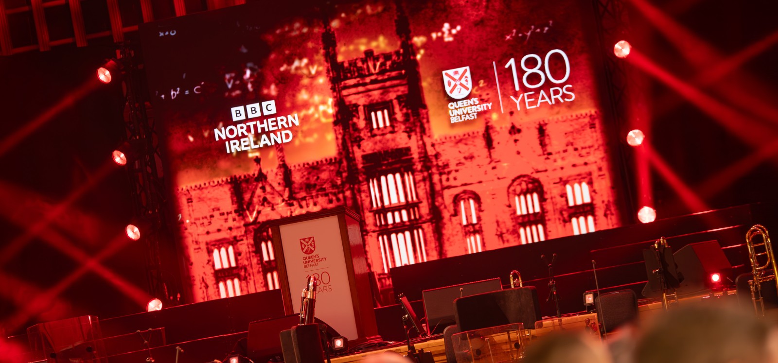 concert stage with presentation screen in red in the background showing logos of BBC Northern Ireland and Queen's University Belfast 180 Years, with picture of the University on the screen