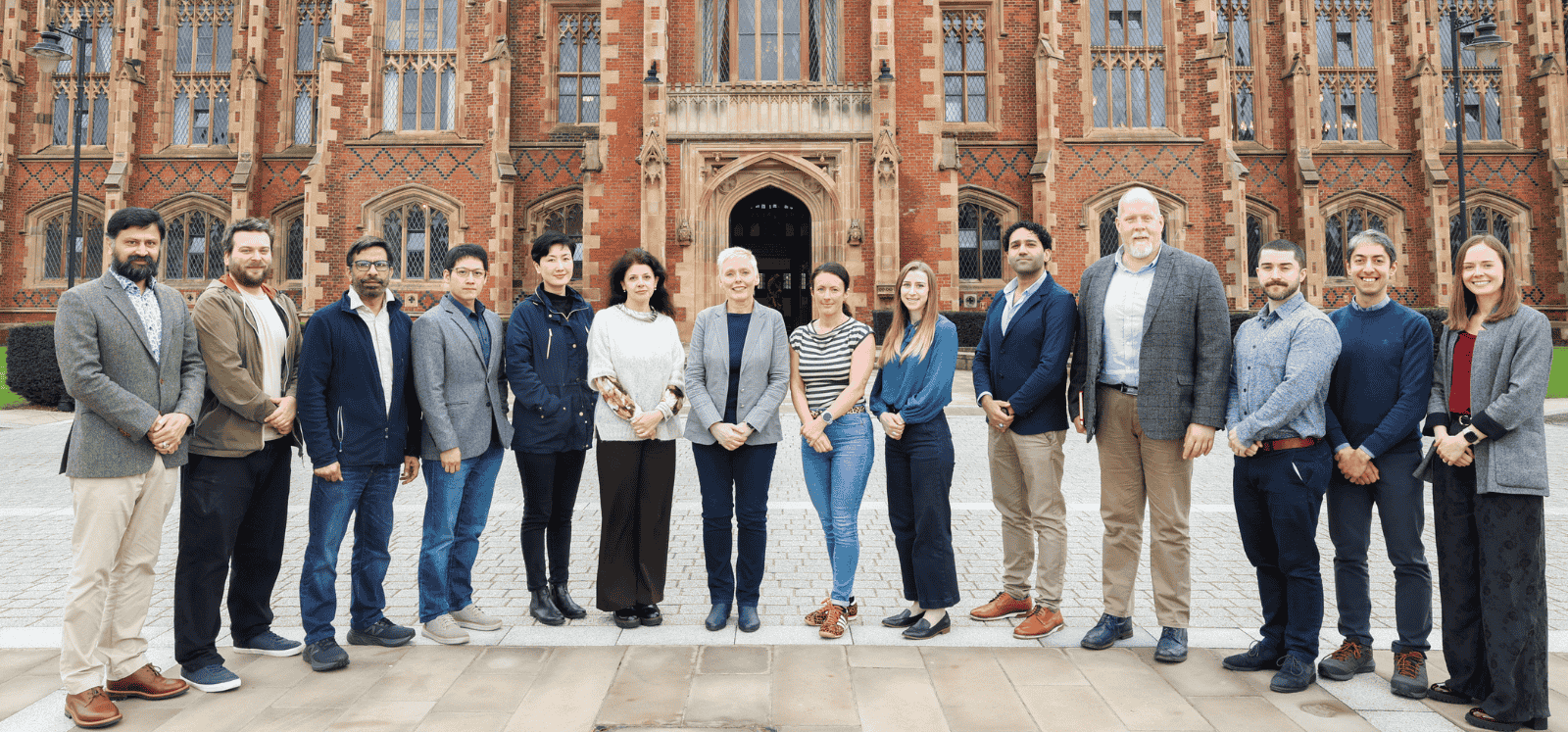 group of 14 men and women standing side by side and posing for the camera on the walkway in front of an old, redbrick building