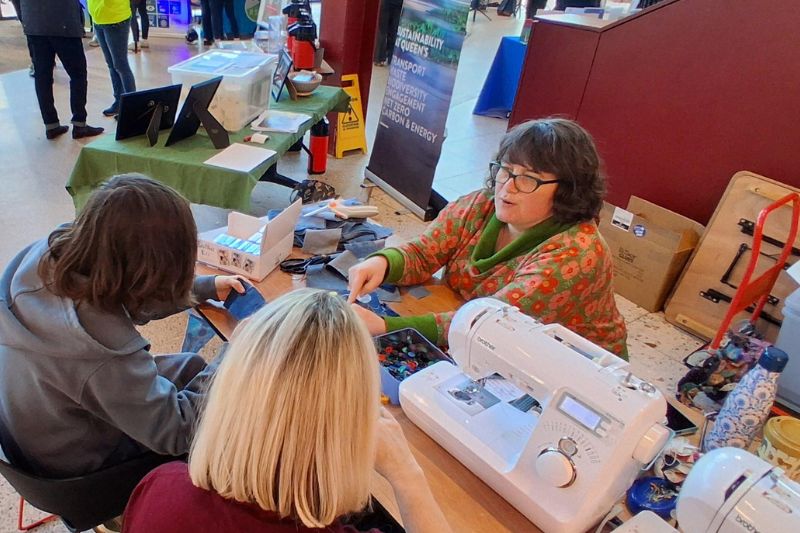 woman sitting at a desk with a sewing kit chatting to two young women at an indoor event