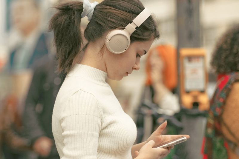 young woman wearing cream or white jumper and headphones using a mobile phone in a public place