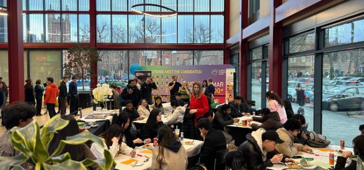 young people seated and in discussion at tables, and other people standing in the background, in the atrium of a modern glass-fronted building