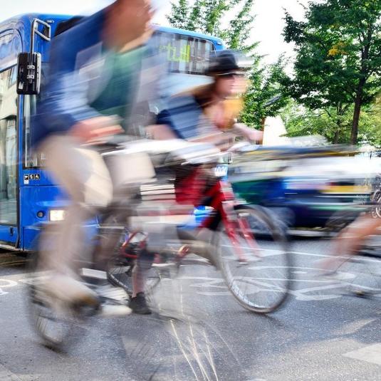 blurry, fast-moving people cycling on a busy road with traffic on a bright day