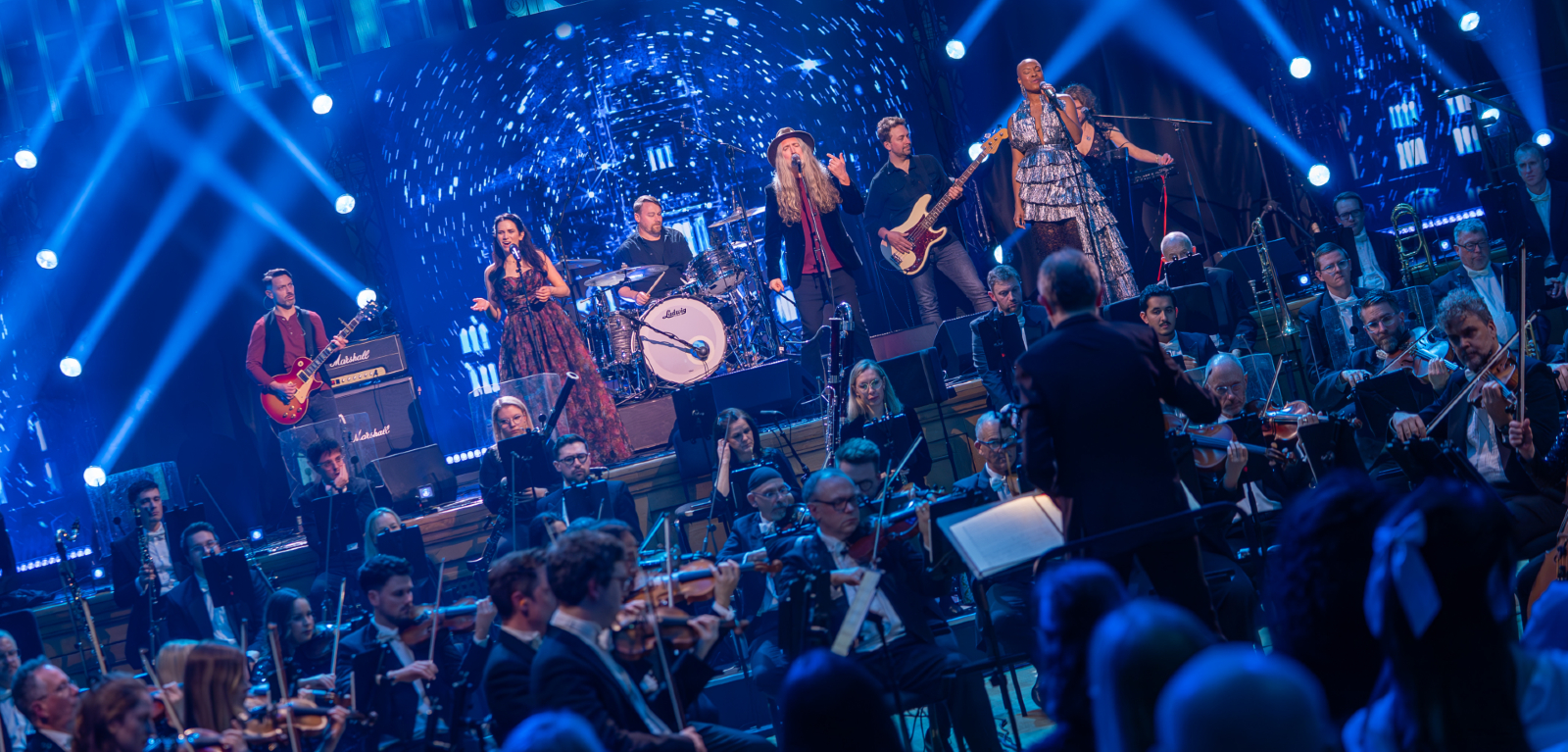 A rock band and singers pictured on a stage performing against a blue starry backdrop with a full orchestra set up on the floor below