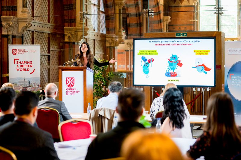 a young woman standing at a podium and presenting to a seated audience in an old but warm-looking redbrick building