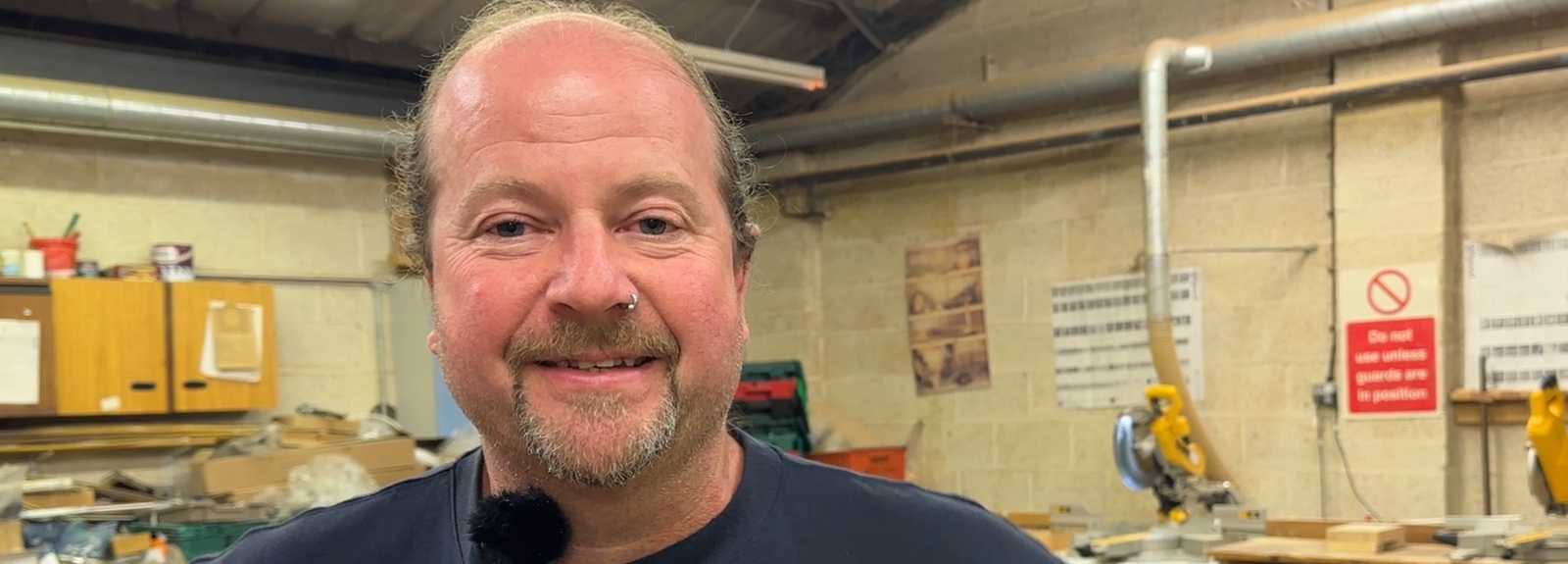 Male member of staff stands in a workshop space with woodworking tools and benches visible in the background.