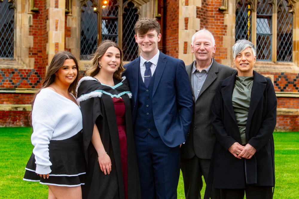 young woman in graduation robe pictured with family and friends outdoors in front of an old redbrick building